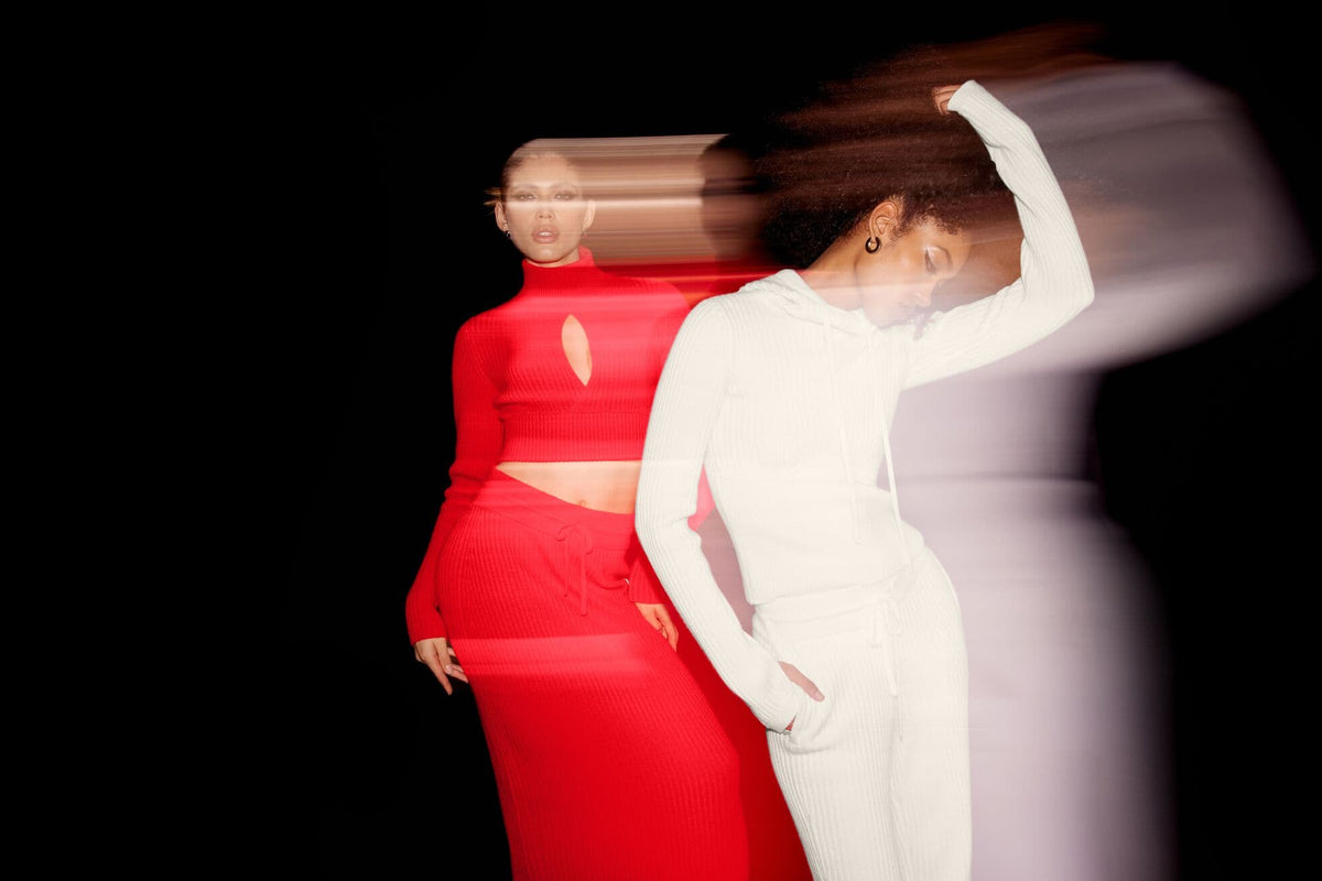 A photo of two women against a black background. One wearing a red cropped cashmere long sleeve top with a floor-length cashmere skirt and the other wearing a white hooded long sleeve cashmere top with wide-leg cashmere pants.  