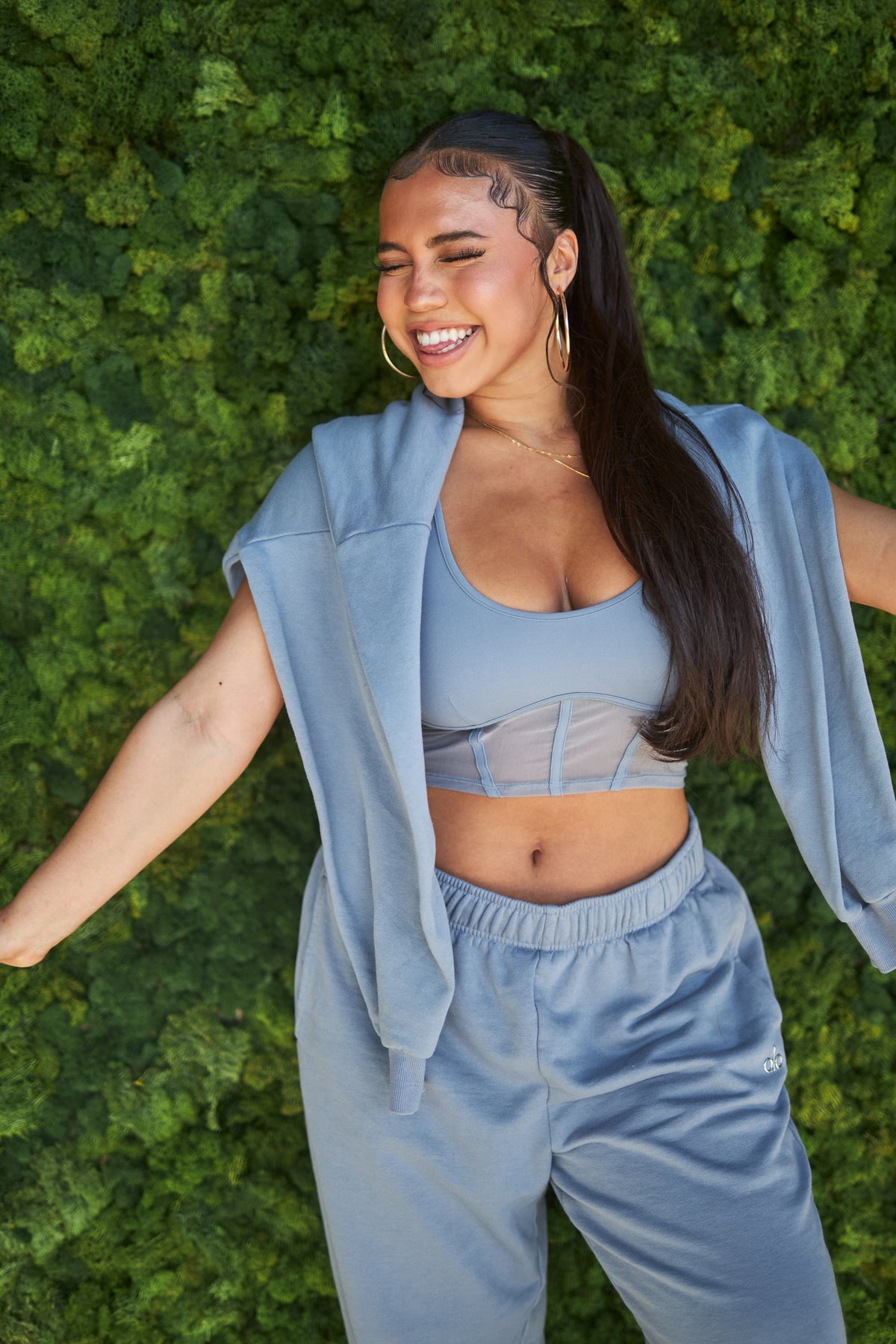 @asiamonetray wearing an Airbrush Mesh Corset Tank in Steel Blue with a matching pair of Accolade Sweat Pants and an Accolade Hoodie draped over her shoulders while posing in front of a wall covered in greenery.  