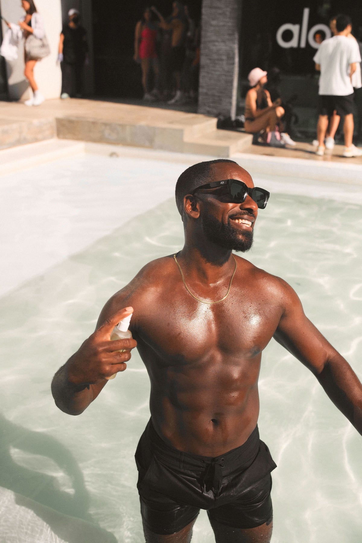 Man wearing Alo shorts standing knee-deep in a swimming pool, spraying his chest with Magnesium Reset Spray 