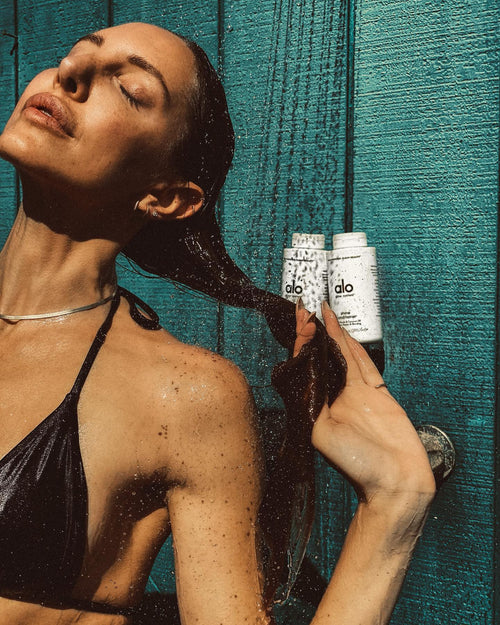 Woman in outdoor shower and black bikini with wet hair in front of Shine Shampoo and Condtitioner